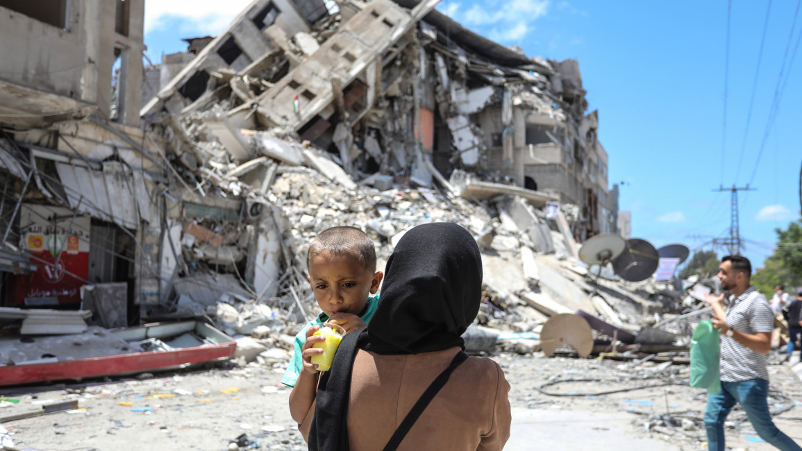 A Palestinian woman carries her baby near the rubble of Al Shorouq tower on 22 May. [Photo by Ahmed Zakot/SOPA Images/LightRocket via Getty Images]