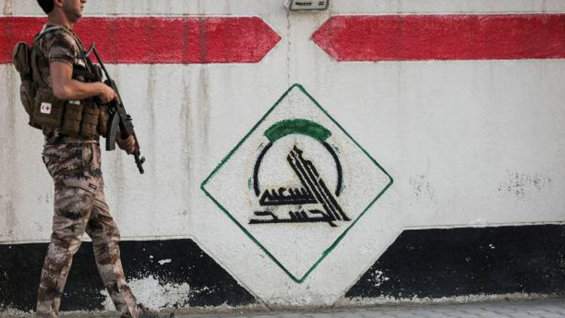 A fighter of Iraq's Popular Mobilisation Forces stands guard next to a wall showing the group's logo outside their headquarters in the capital Baghdad on June 13, 2021. [Photo by AHMAD AL-Rubaye/AFP via Getty Images]