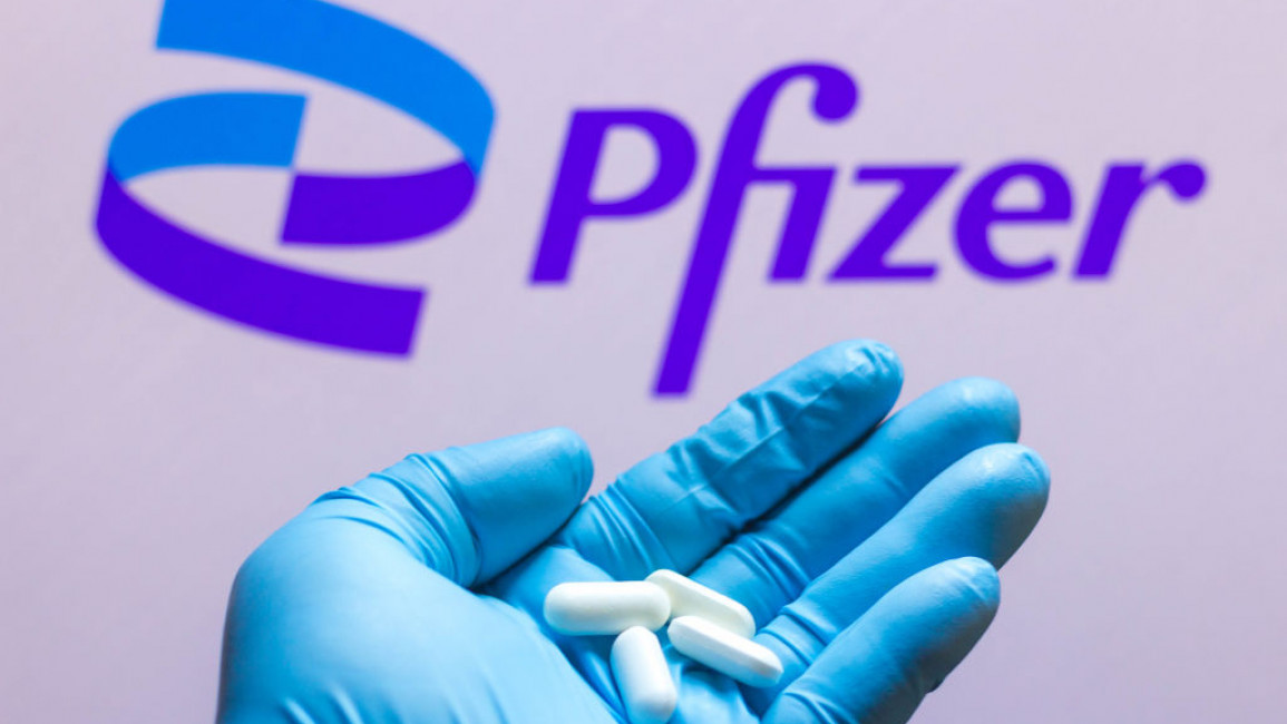 A person's hand inside a blue medical glove holding four white pills in front of a Pfizer logo