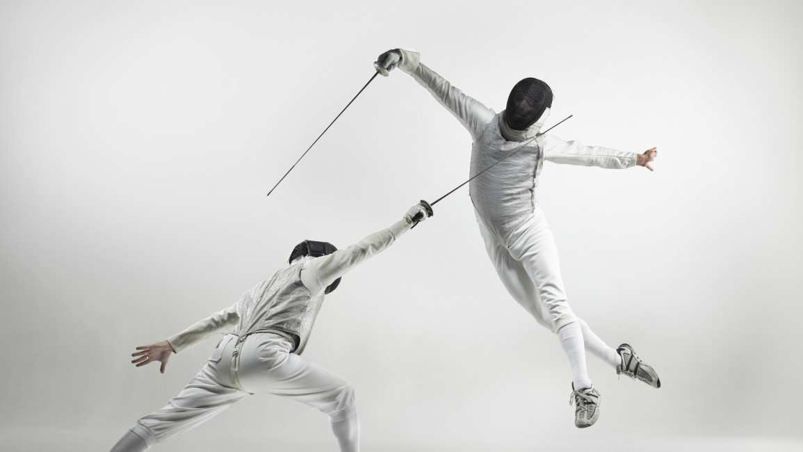 There are two fencers competing. The one on the left is high in the air, jumping down on the second, who is lunging up at the first