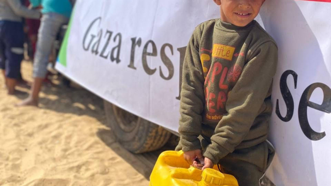 A child beneficiary of Molham's aid project in Gaza stands beneath an banner holding a water canister. [Image provided courtesy of Molham]