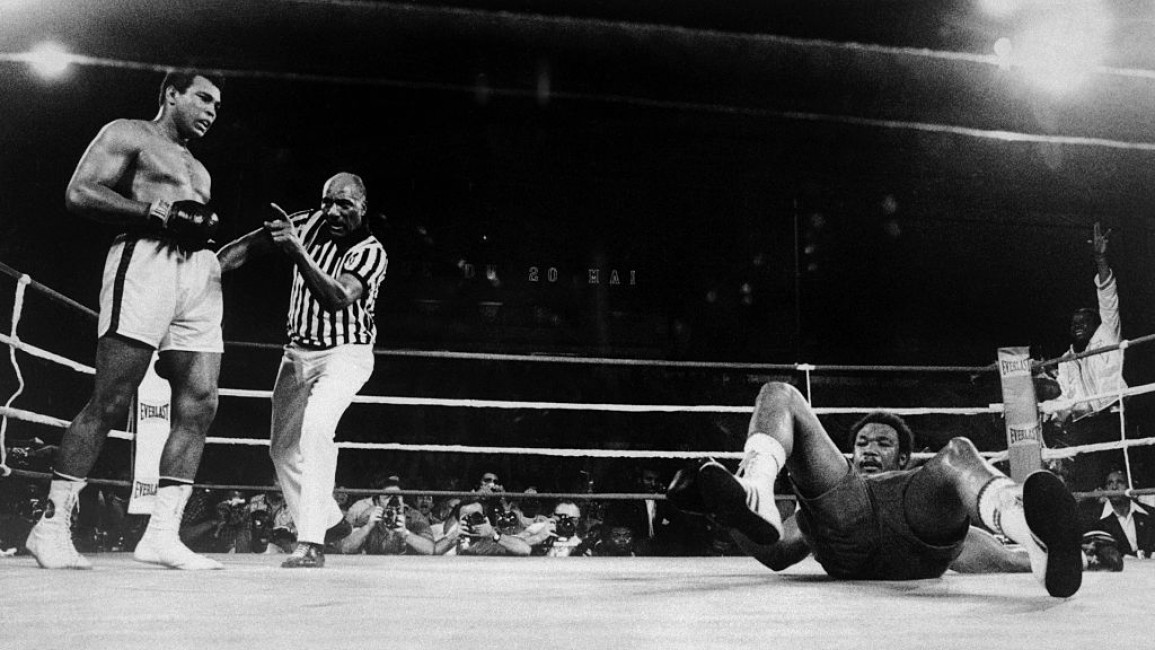 A photo showing George Foreman (right) on his back, with Muhammad Ali standing up (far left).